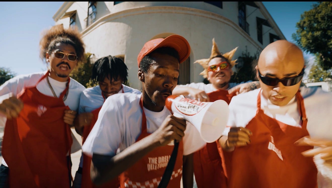 Crew in red Deli Tour aprons walking a sunlit street, one leading a chant through a megaphone